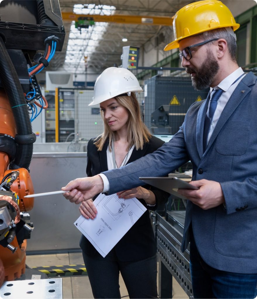 Eine Dame und ein Herr in Businessmontur und mit Helmen diskutieren Arbeitssicherheit an einer Maschine